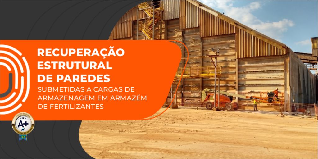 Fachada lateral de um grande armazém de fertilizantes em estrutura metálica e fechamento em placas de concreto pré-moldado, exibindo operários e maquinário articulado laranja realizando serviços de manutenção e recuperação estrutural na parte externa da edificação