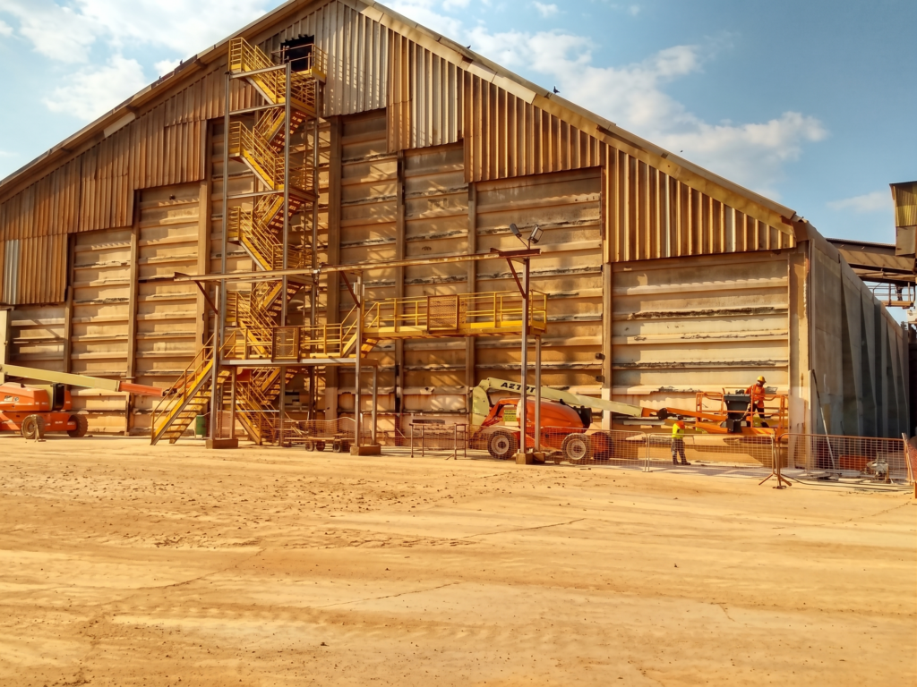 Vista externa da fachada lateral de um armazém de fertilizantes estruturado com pilares pré-moldados e fechamento em placas de concreto, evidenciando marcas de fissuras horizontais nos painéis e equipes técnicas operando plataformas elevatórias articuladas articuladas para os serviços de engenharia estrutural