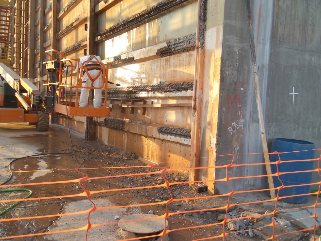 Operário equipado com cinturão de segurança sobre uma plataforma elevatória realiza o hidrojateamento de alta pressão nas mísulas e placas de concreto de um armazém de fertilizantes, removendo o concreto degradado e expondo as armaduras de aço oxidadas para o processo de tratamento estrutural