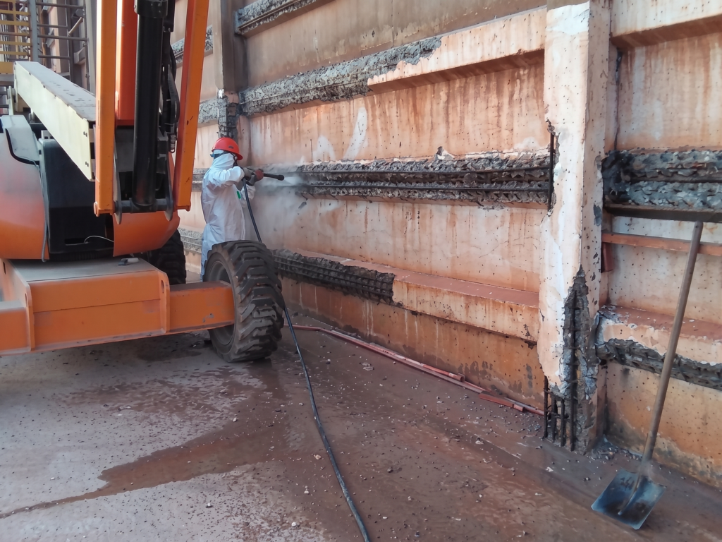 Operário equipado com cinturão de segurança sobre uma plataforma elevatória realiza o hidrojateamento de alta pressão nas mísulas e placas de concreto de um armazém de fertilizantes, removendo o concreto degradado e expondo as armaduras de aço oxidadas para o processo de tratamento estrutural