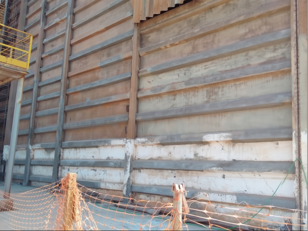 Placas pré-moldadas de concreto em parede estrutural de armazém com marcas escuras de umidade e fissuras horizontais evidentes na base dos painéis, protegida na parte inferior por uma tela de sinalização laranja