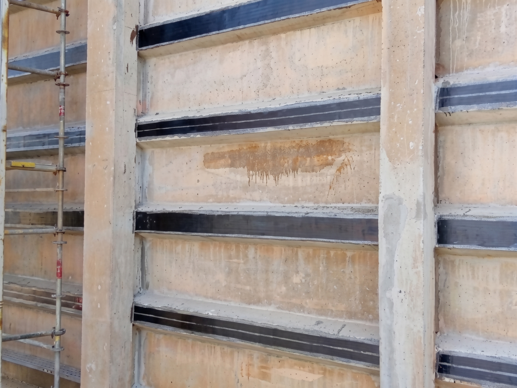Placas pré-moldadas de concreto instaladas entre pilares estruturais, exibindo faixas horizontais pretas de fibra de carbono coladas e aplicadas sobre a região das mísulas para o reforço estrutural, com parte de uma estrutura de andaime metálico visível à esquerda