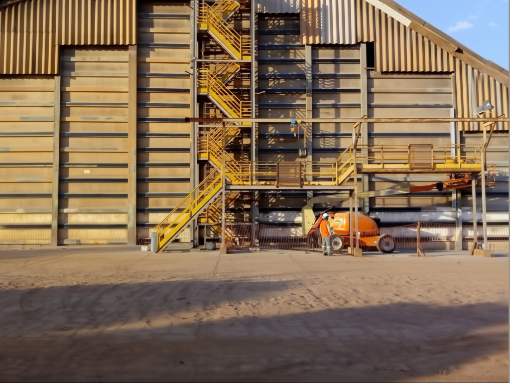 Fachada de armazém de fertilizantes mostrando detalhe da escada metálica amarela de acesso e operário de capacete branco posicionado ao lado de uma plataforma elevatória articulada laranja durante a execução das obras de engenharia